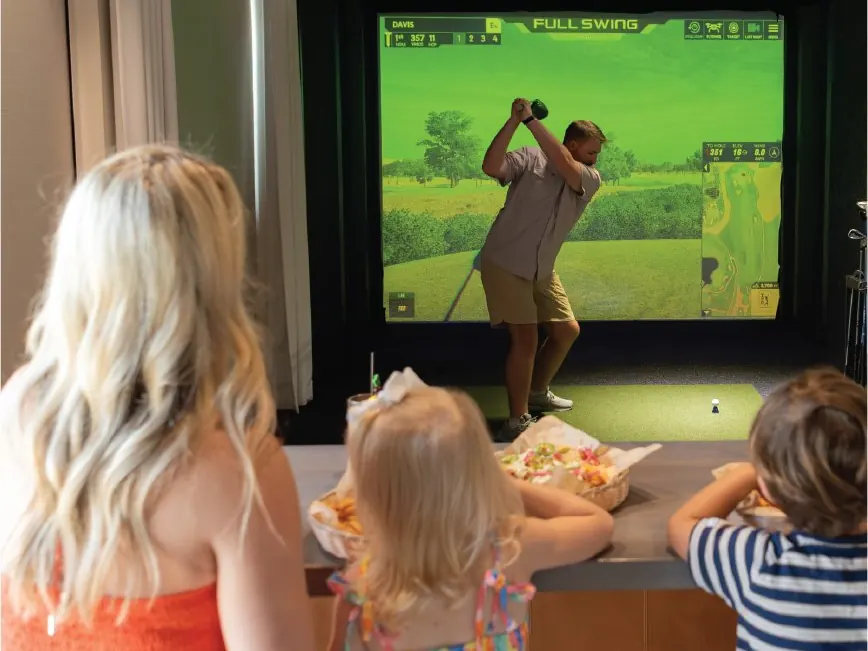 A golfer mid-swing in a simulator bay at Tee Off at The Wharf while a family watches from the counter