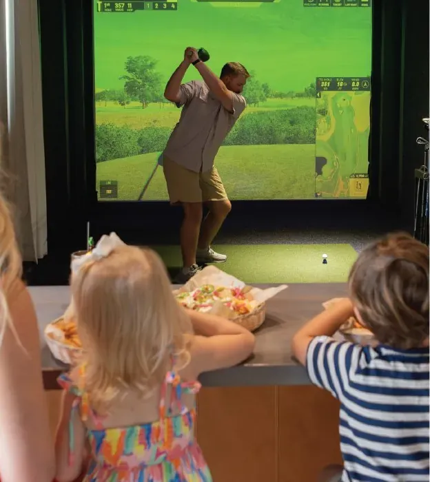 A family watching a golfer swing in a simulator bay at Tee Off at The Wharf