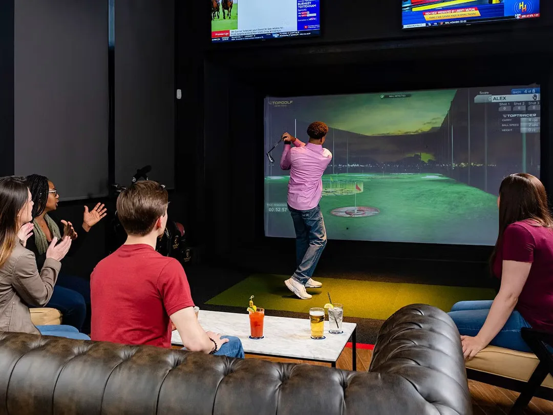 A group watching a golfer take a swing in a simulator bay at Tee Off at The Wharf